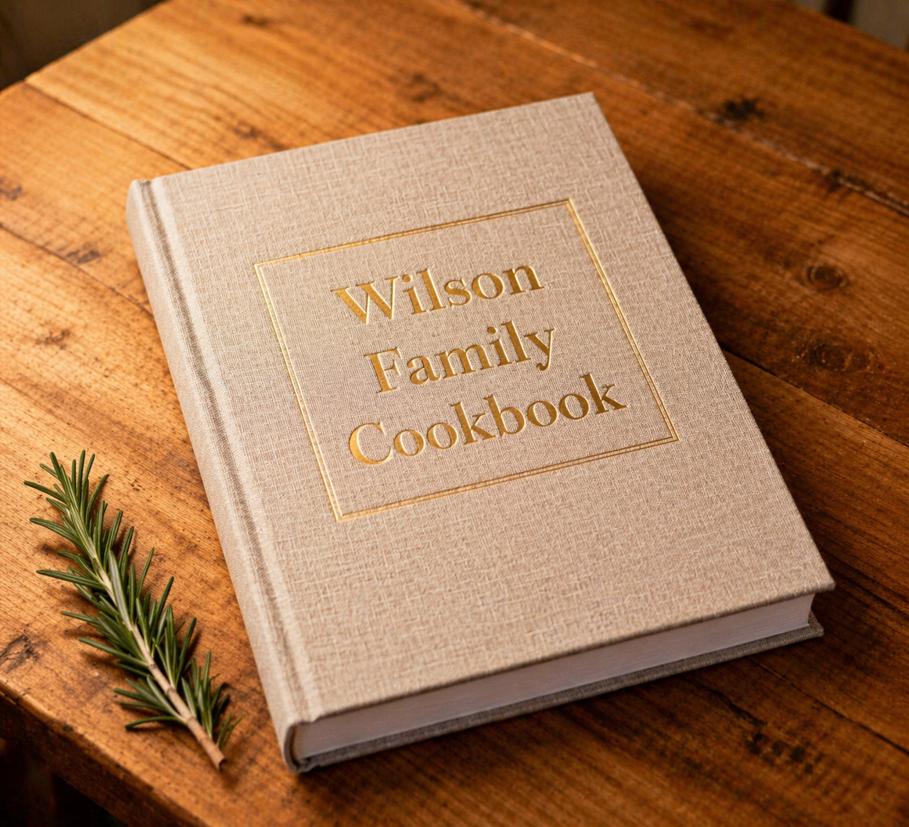 A linen-bound printed cookbook resting on a wooden table with a sprig of rosemary